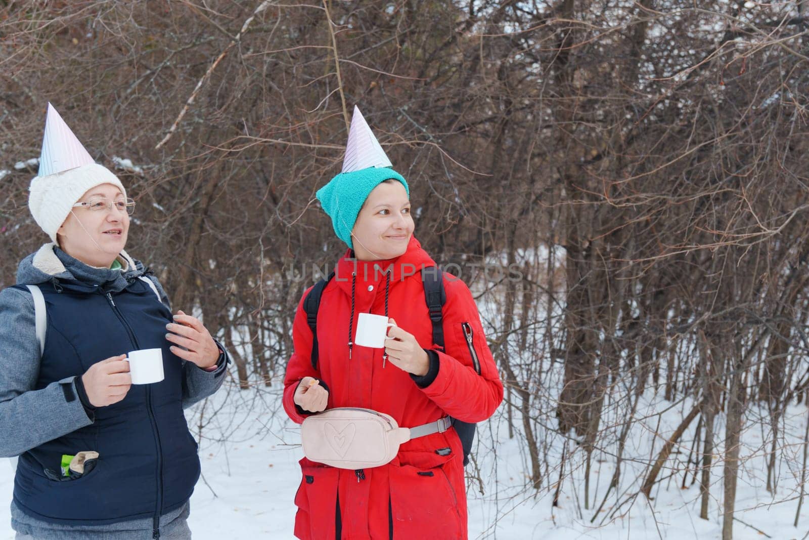 Two friends in festive party hats enjoy warm beverages while standing in a snowy area.