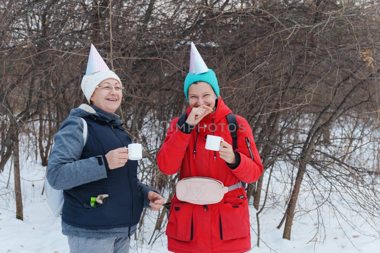 Two friends in festive party hats enjoy warm beverages while standing in a snowy area.