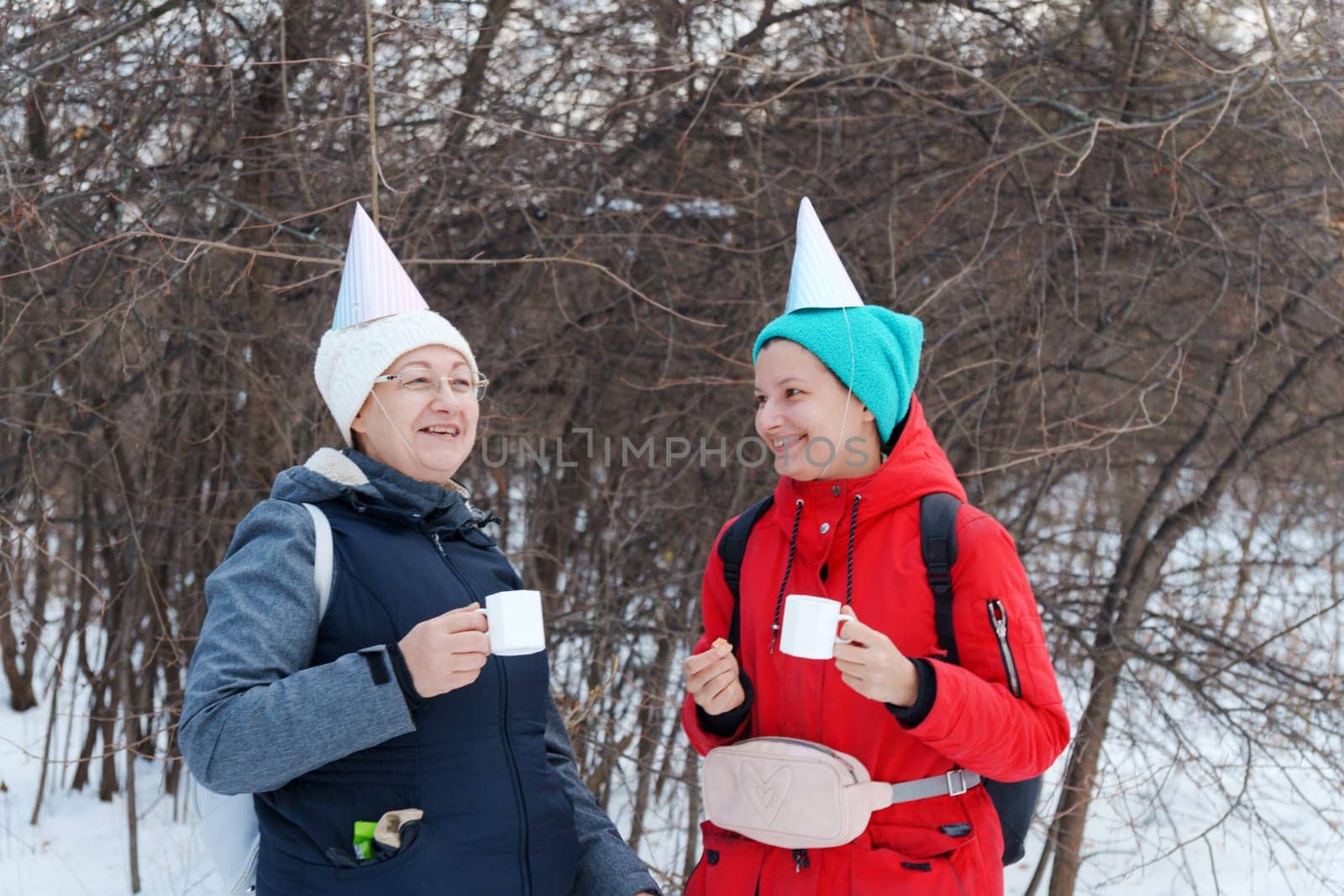 Two friends in festive party hats enjoy warm beverages while standing in a snowy area.