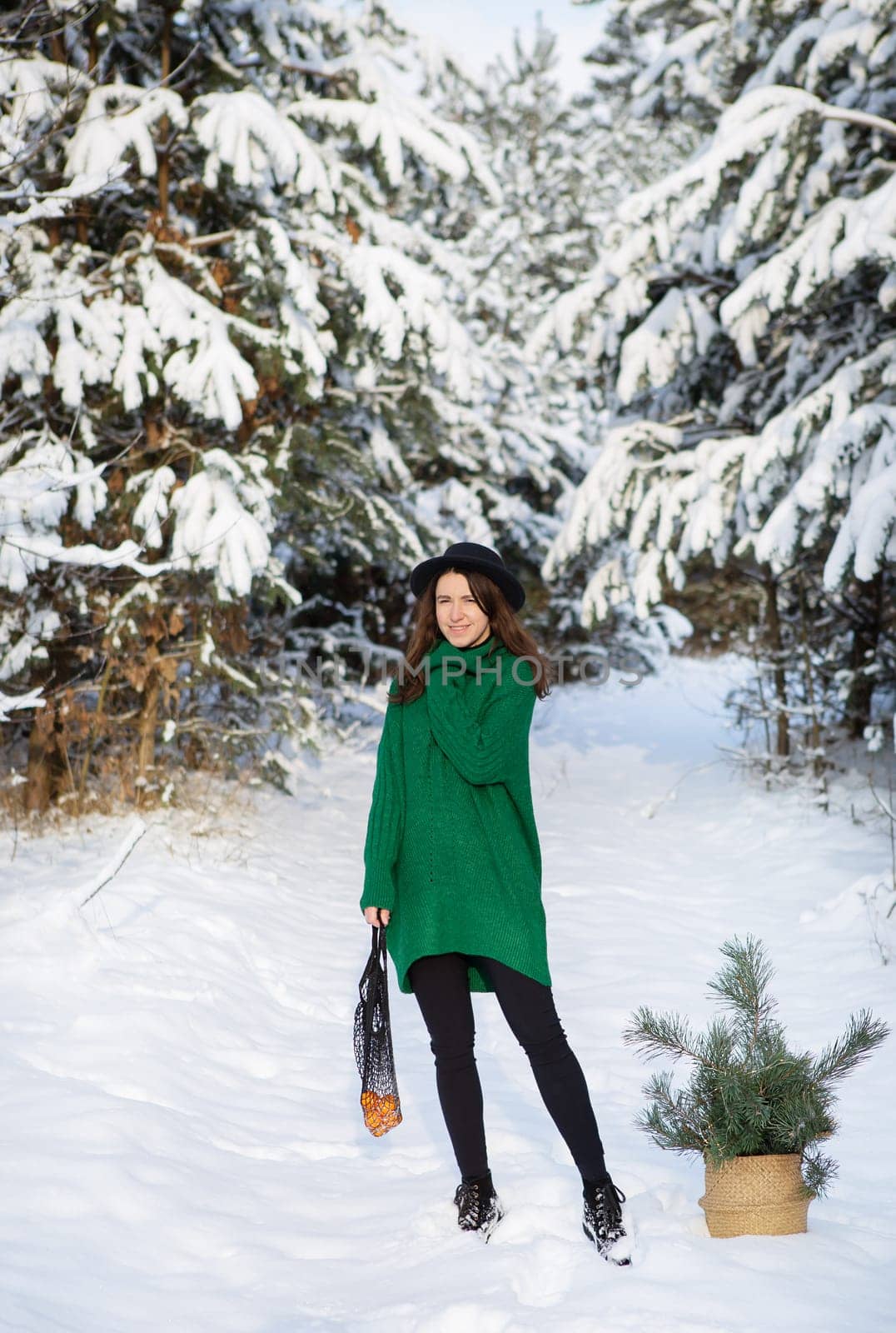 A young beautiful girl in a green sweater and hat stands in the winter forest with a basket, with pine branches and a string bag with tangerines.