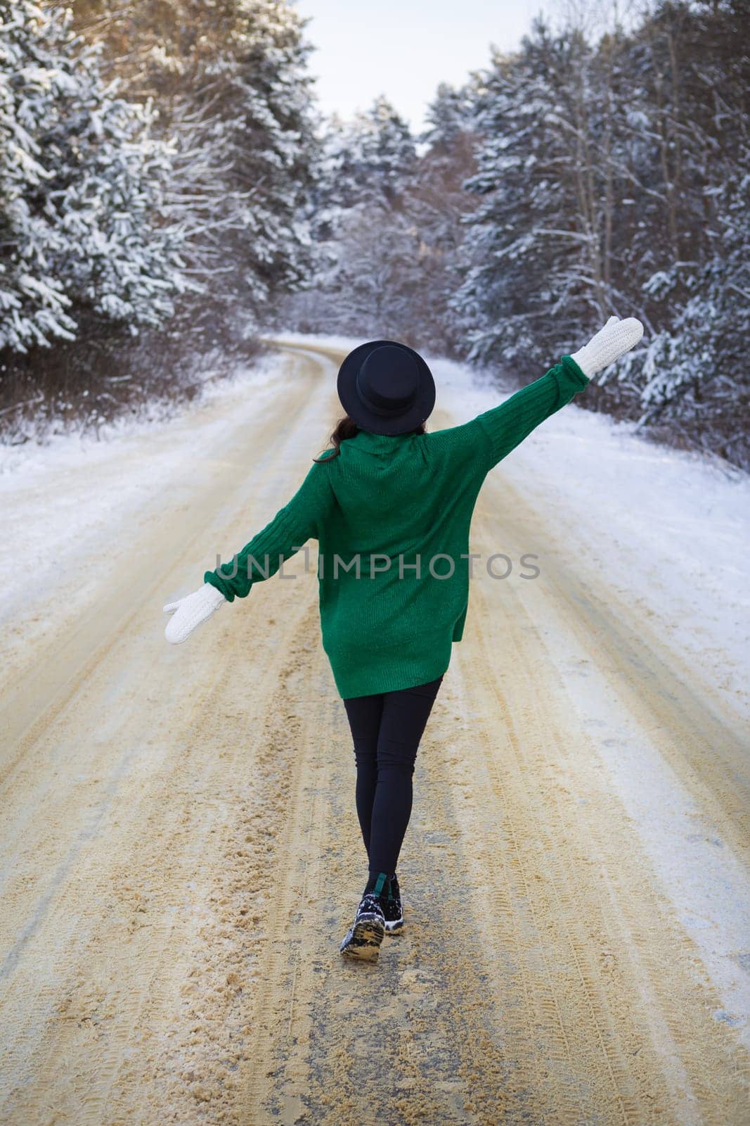 A young girl in a green sweater and hat walks in the middle of a snowy road in a thick pine forest. Freezing day