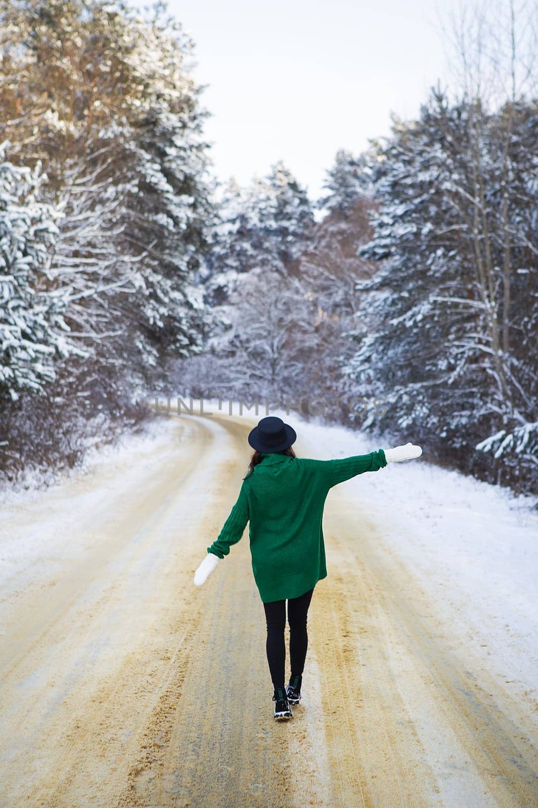 A young girl in a green sweater and hat walks in the middle of a snowy road in a thick pine forest. Freezing day