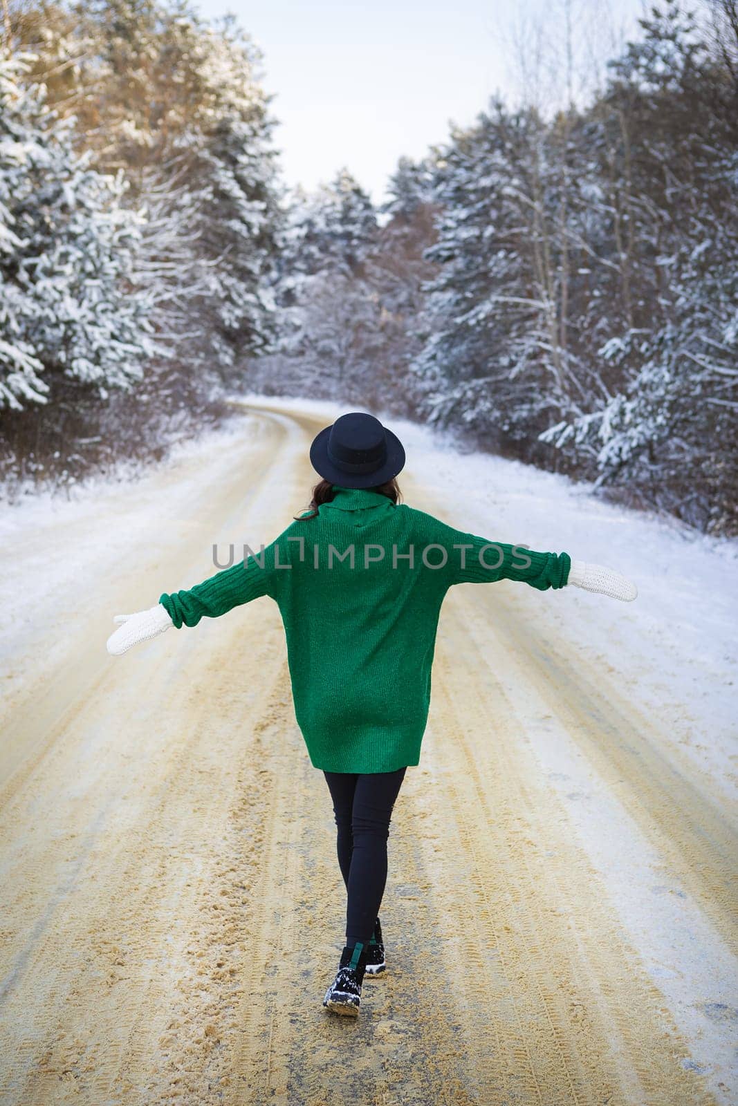 A young girl in a green sweater and hat walks in the middle of a snowy road in a thick pine forest. Freezing day