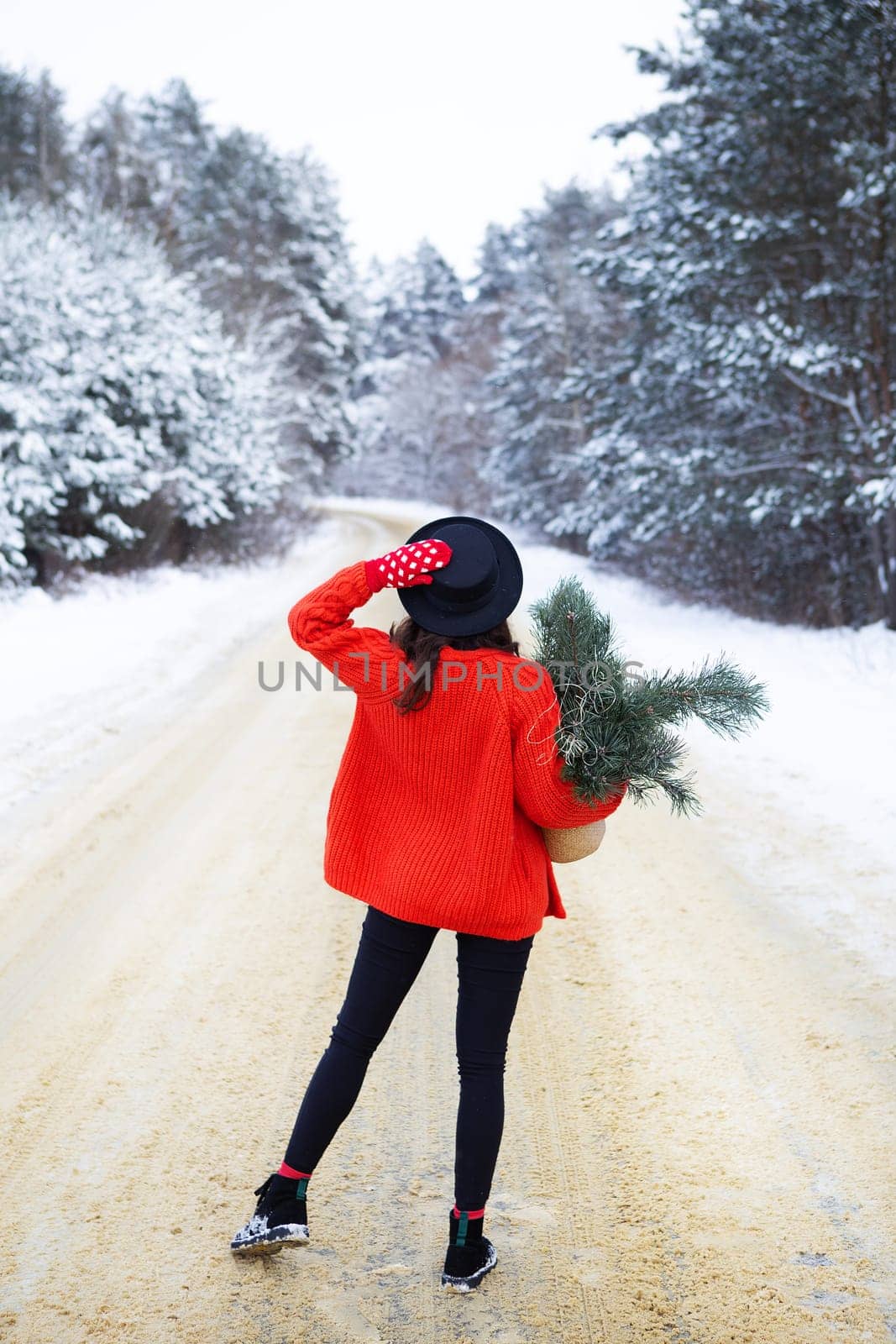 A girl in a red sweater and hat stands in the middle of a snowy road in a forest with pine branches. Weekend trip
