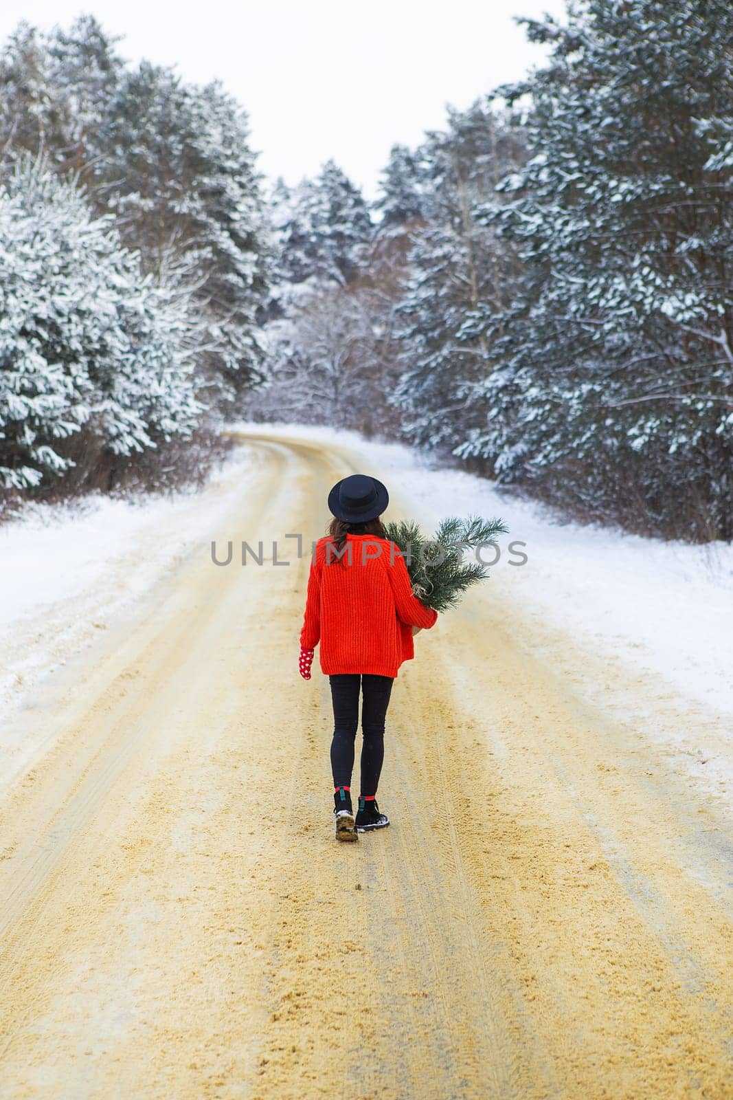 A girl in a red sweater and hat stands in the middle of a snowy road in a forest with pine branches. Weekend trip