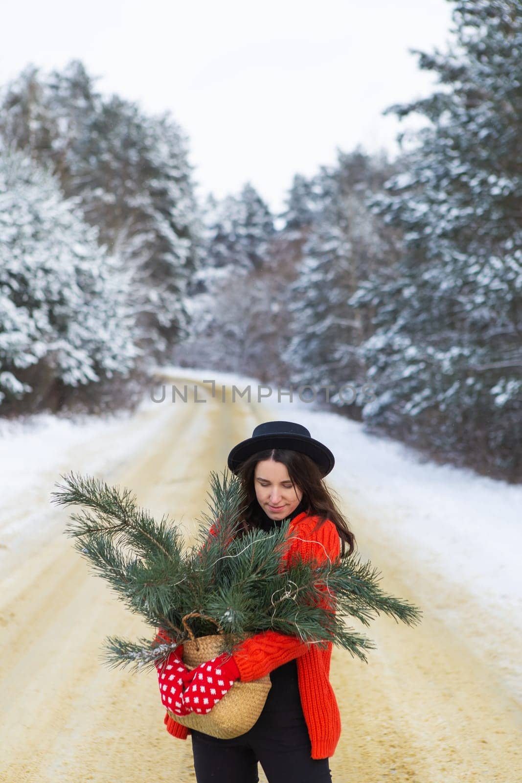 A girl in a red sweater and hat, in red mittens holds a basket with pine branches in her hands stands in the middle of a snow-covered road in a forest with pine branches