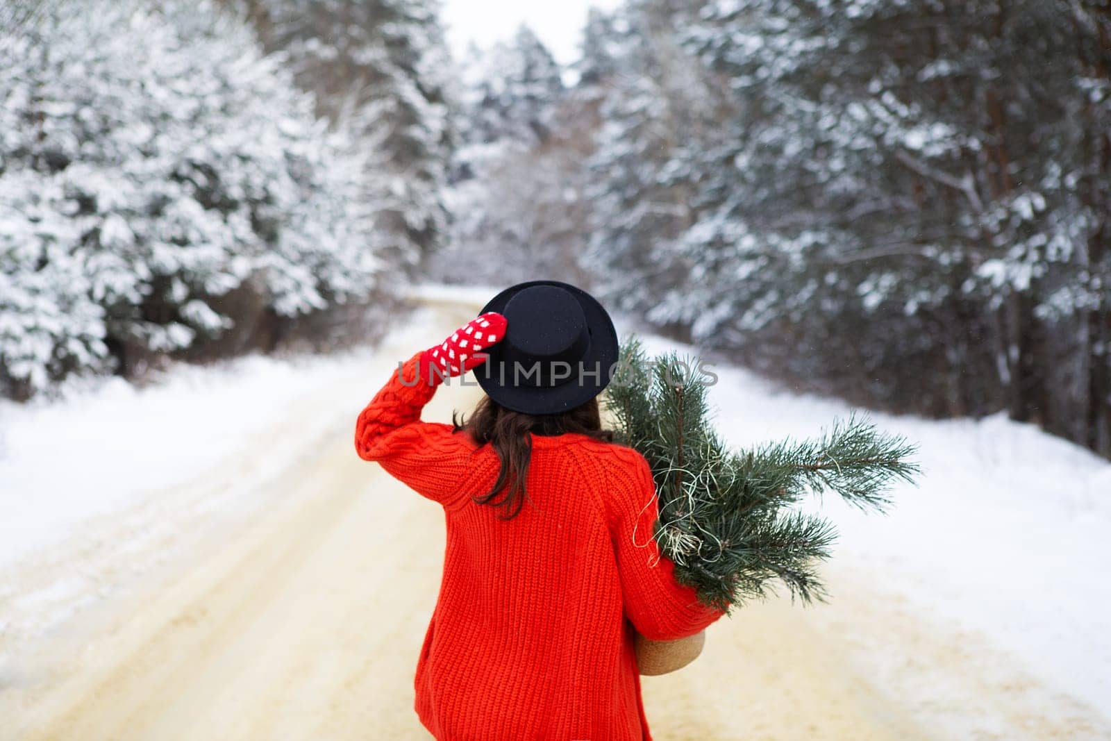 A girl in a red sweater and hat stands in the middle of a snowy road in a forest with pine branches. Weekend trip
