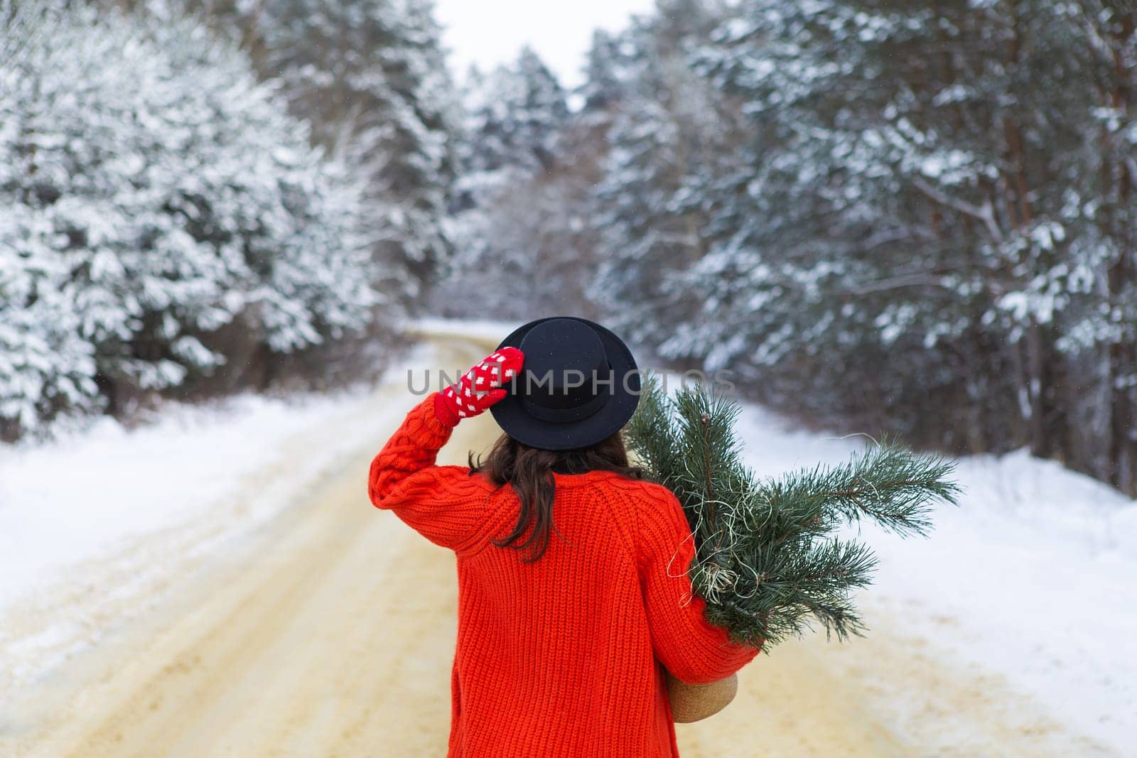 A girl in a red sweater and hat, red mittens stands in the middle of a snow-covered road in a forest with pine branches. Weekend trip