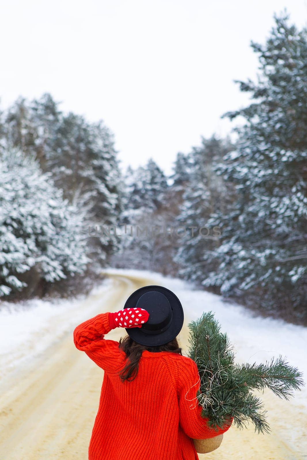 A girl in a red sweater and hat stands in the middle of a snowy road in a forest with pine branches. Weekend trip
