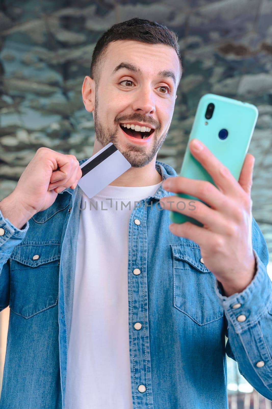 Happy young man using his smartphone while holding a credit card. He smiles with excitement, standing outside a glass building, enjoying an online shopping or payment experience. Vertical