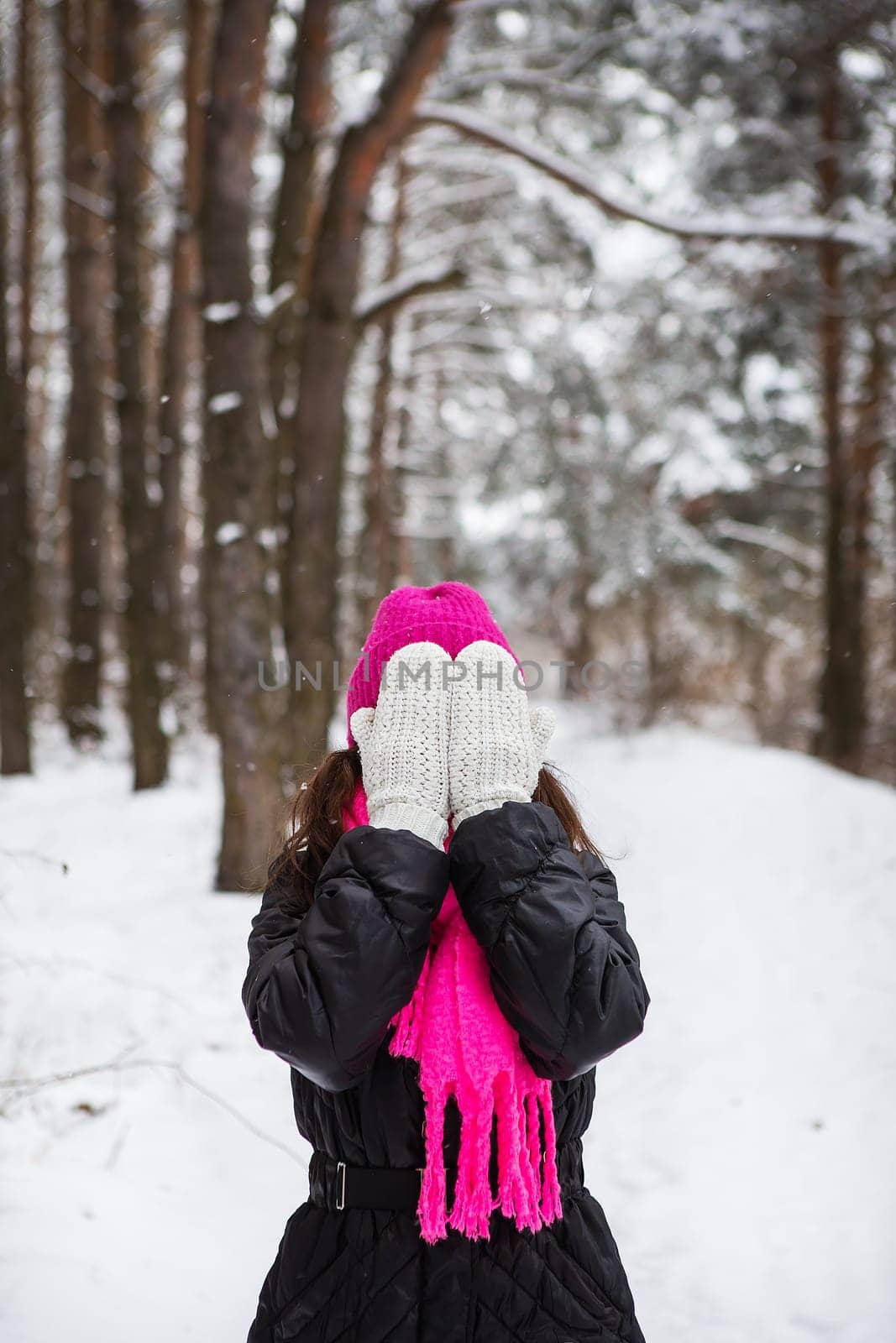 A girl in a pink scarf and hat stands in the middle of a pine forest and covers her face with white mittens. Beautiful pine forest on a frosty day