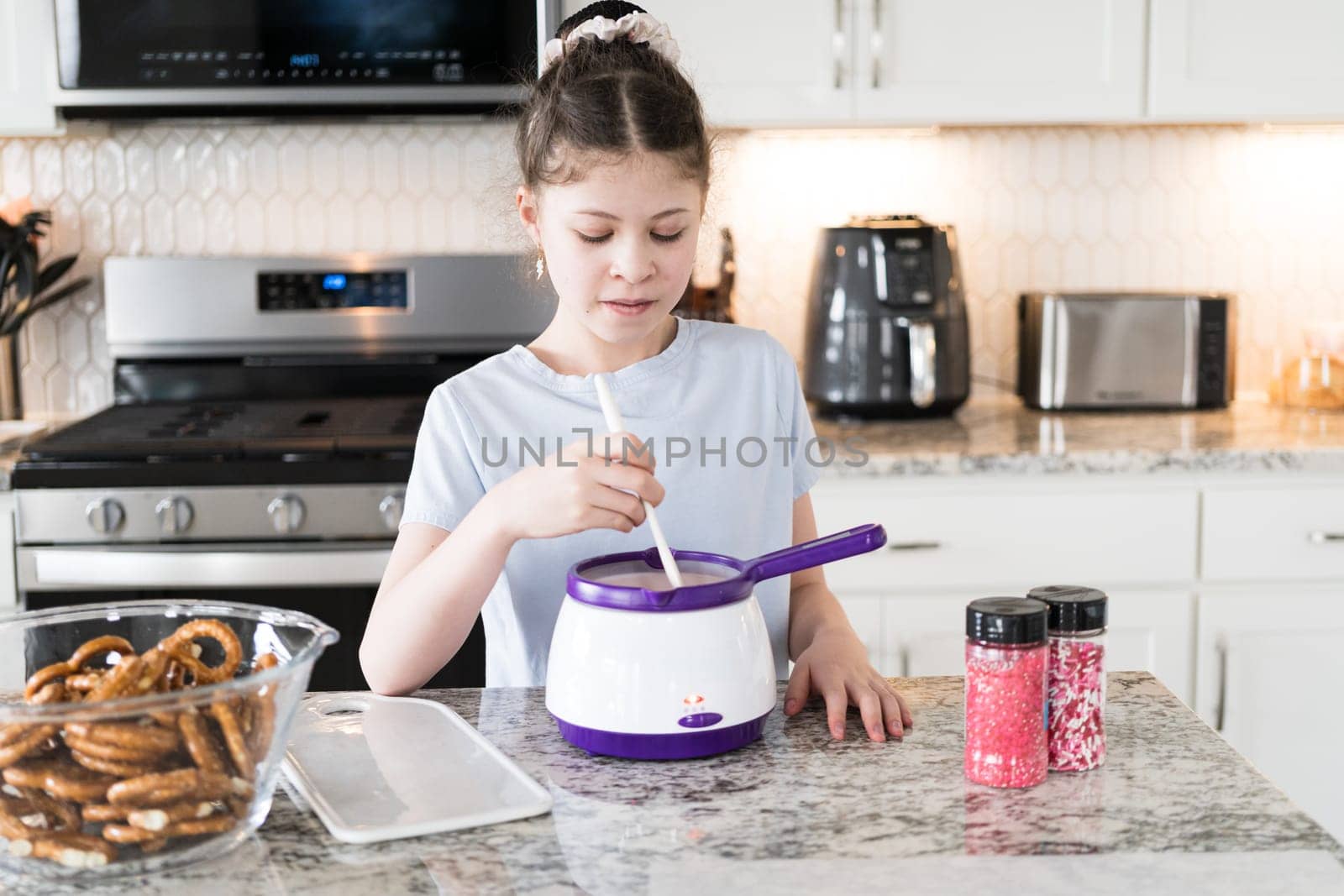 With focused attention, this budding culinary artist dips pretzels into a pot of melted chocolate, creating sweet delights in the warmth of a well-lit home kitchen.