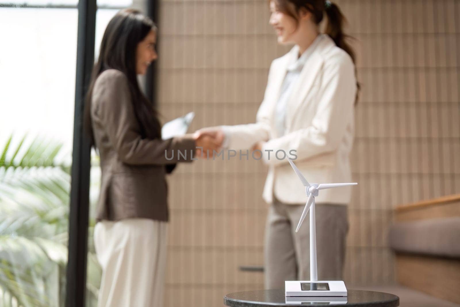 In a modern office setting, two businesswomen shake hands, symbolizing their commitment to sustainability while featuring a model of a wind turbine,