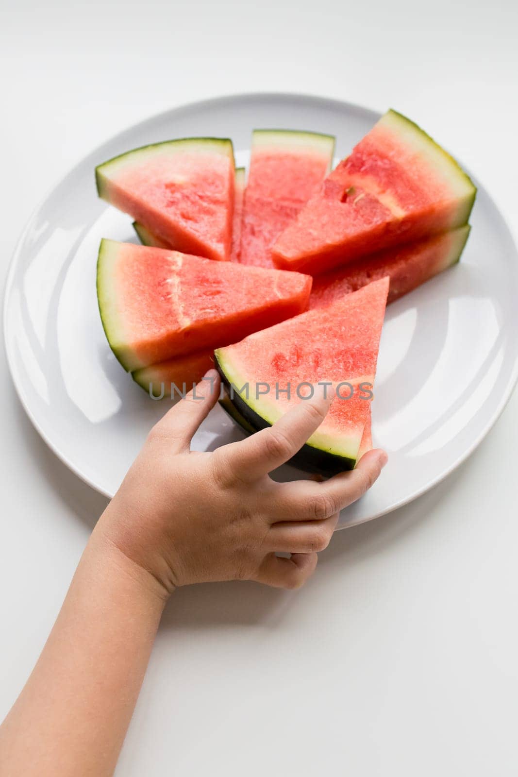 Child hand reaching for fresh watermelon slices on white plate.