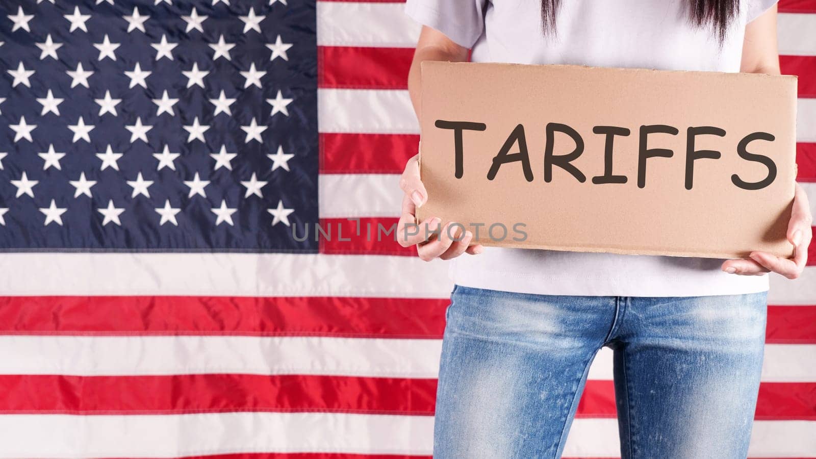 A young woman stands before an American flag, holding a sign that says tariffs in black marker. She wears a white shirt and blue jeans. The photo is from a low angle, concealing her face.