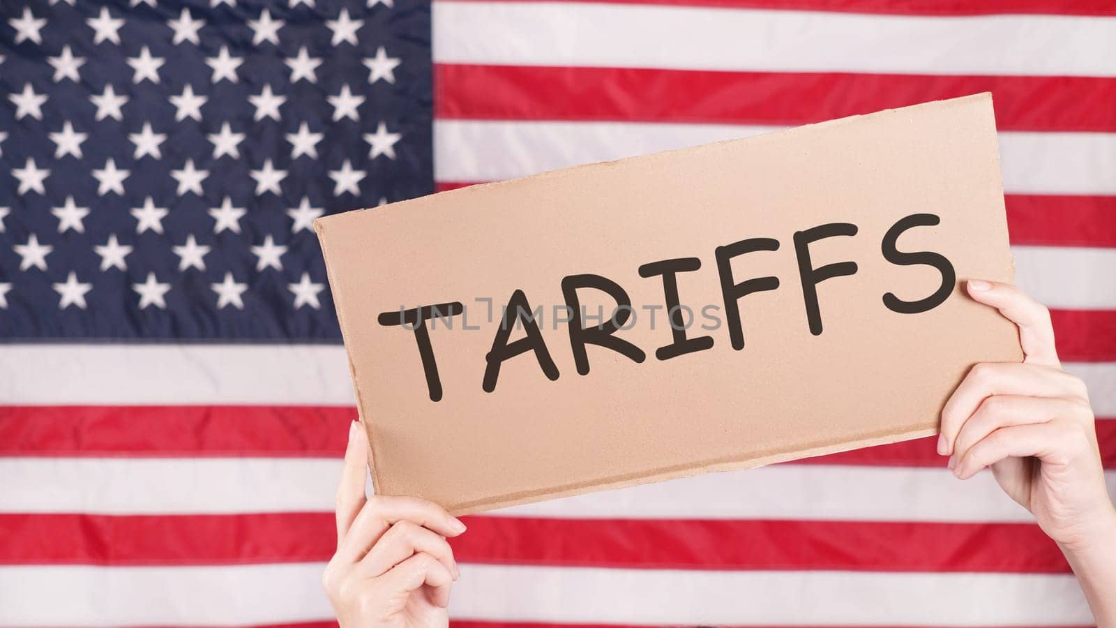 A cardboard sign with the word Tariffs written on it is held up in front of an American flag. The sign is held by a pair of hands and the background is out of focus.