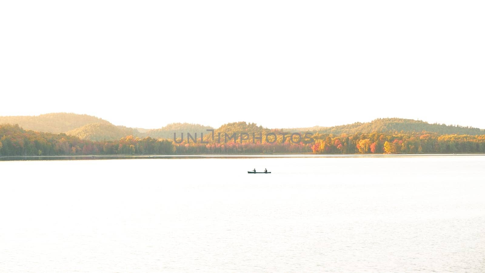 The image shows a small boat on a calm lake with a mountain range in the background. The boat is rowed by two people and the sun is rising or setting, casting a golden glow over the scene.