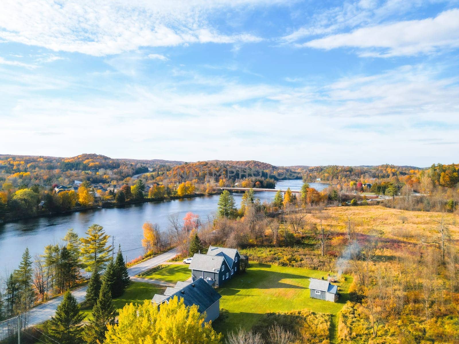 A scenic rural landscape showcases a flowing river, vibrant autumn trees, and a cozy house by the water. The clear blue sky with white clouds enhances the peaceful atmosphere.