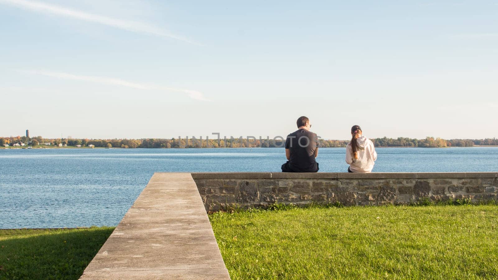 A young couple is sitting on a stone wall overlooking a tranquil lake. They are gazing out at the stunning view and enjoying the peace and quiet, surrounded by nature s beauty and serenity.