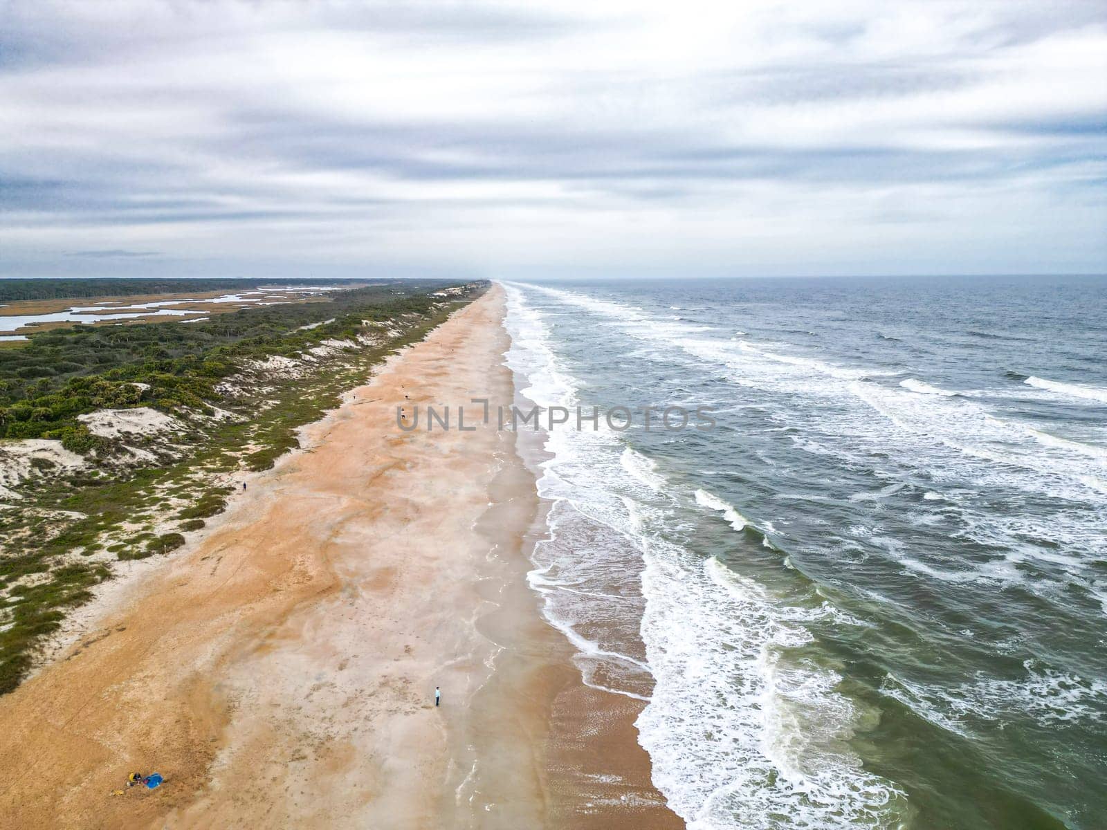 The beach is wide and sandy with a gentle slope into the ocean. The water is clear and blue-green. The sky is blue with scattered clouds. There are dunes covered with sea oats behind the beach.