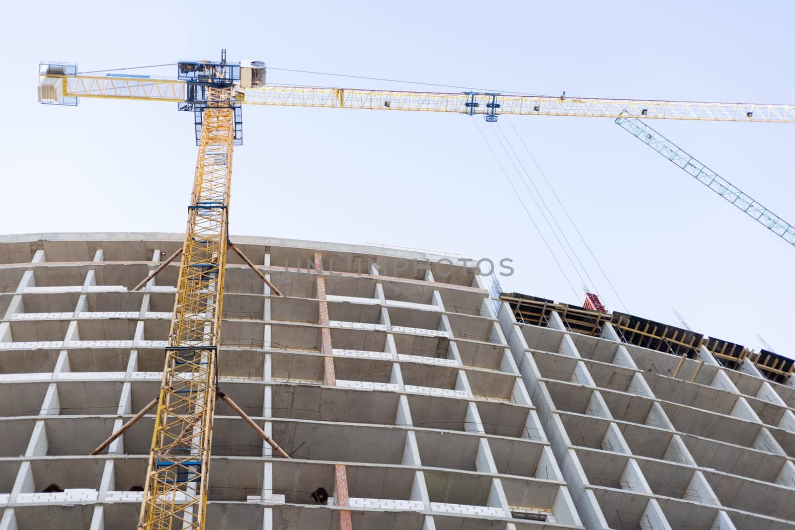 Concrete frame of a multi-story building under construction with large yellow and blue cranes operating against a clear sky background