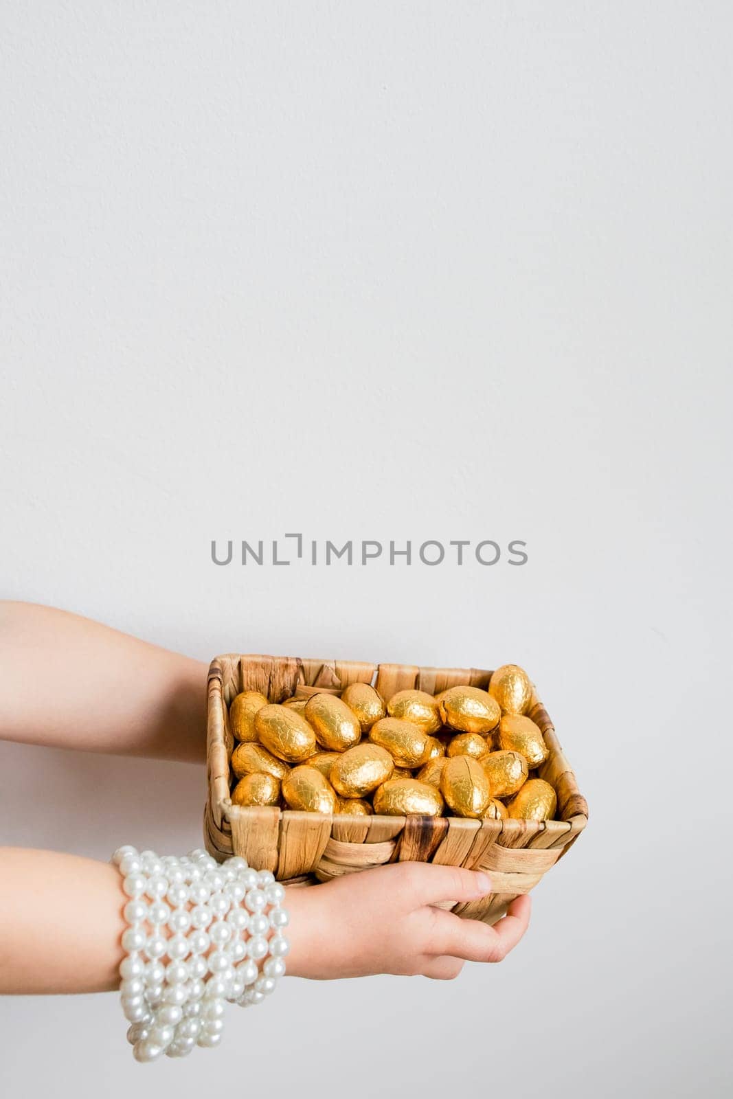 Child holding basket of gold wrapped chocolate eggs against white background.