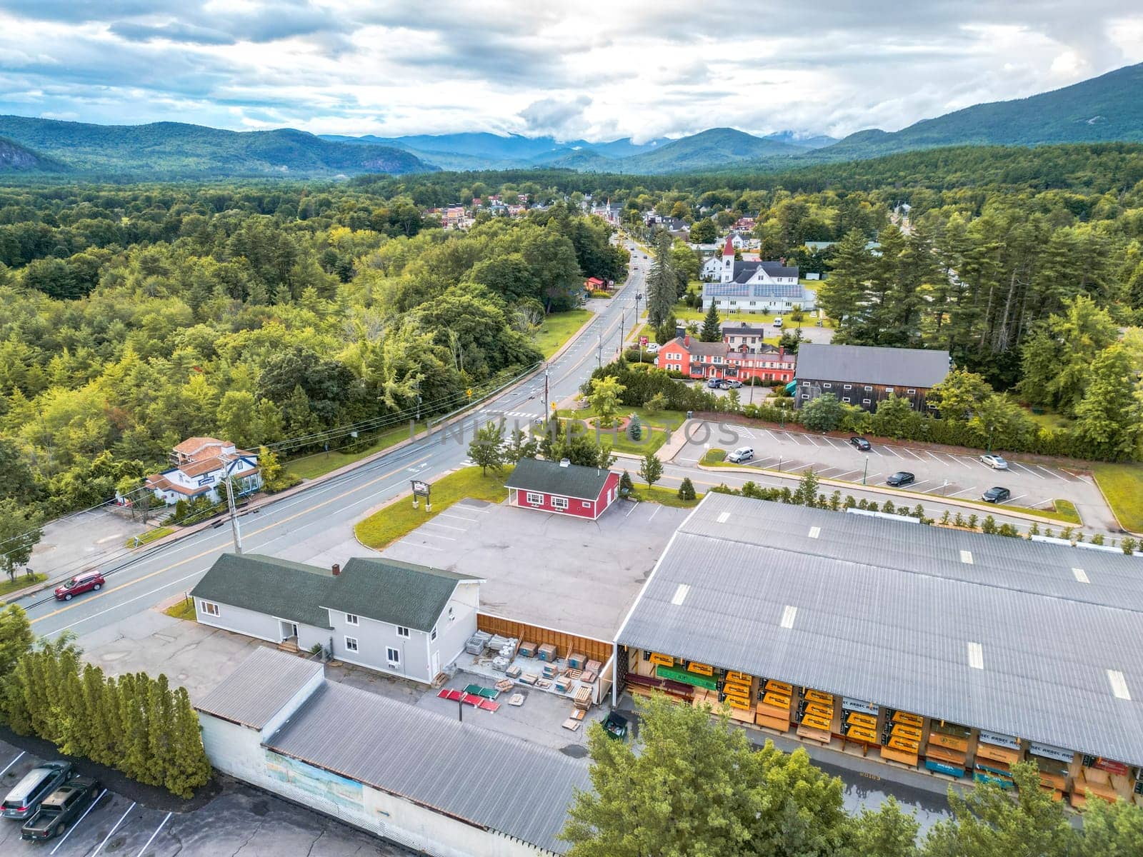 This stunning aerial view captures a charming New England town nestled among majestic mountains. Lush green forests surround the area, with a clear blue sky and fluffy clouds enhancing the beauty.