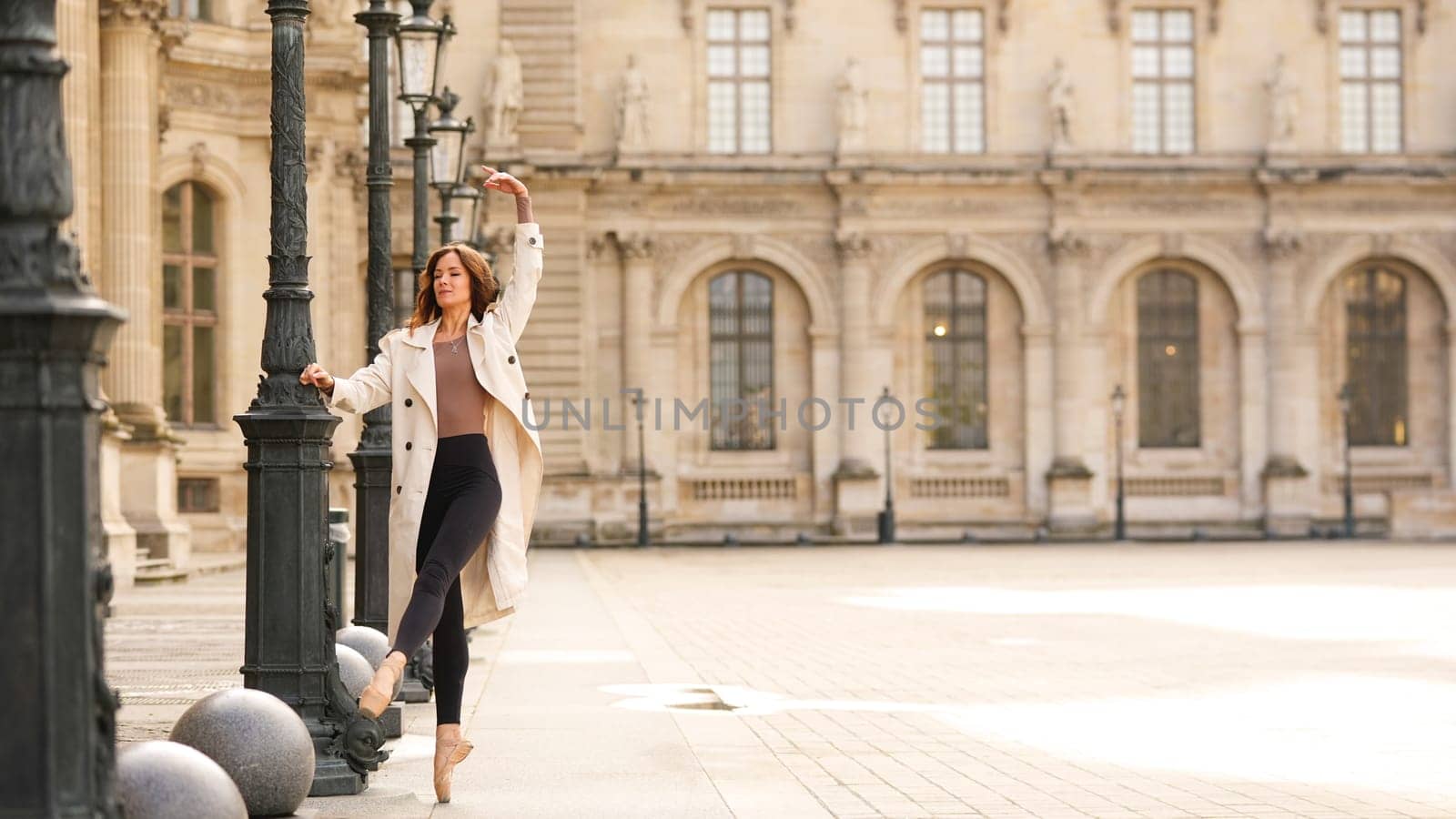 PARIS, FRANCE 11 APRIL 2025 A ballet dancer in a beige trench coat and pointe shoes performs a graceful movement near the historic Louvre Museum