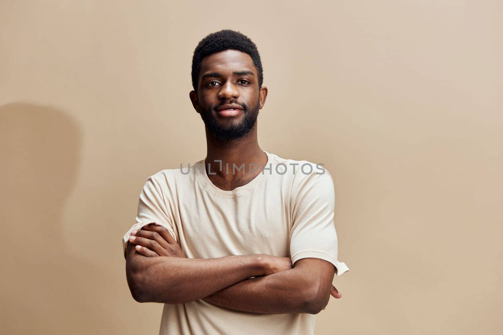 A confident young African man wearing a beige t shirt stands with his arms crossed against a warm background, exuding self assurance and calmness