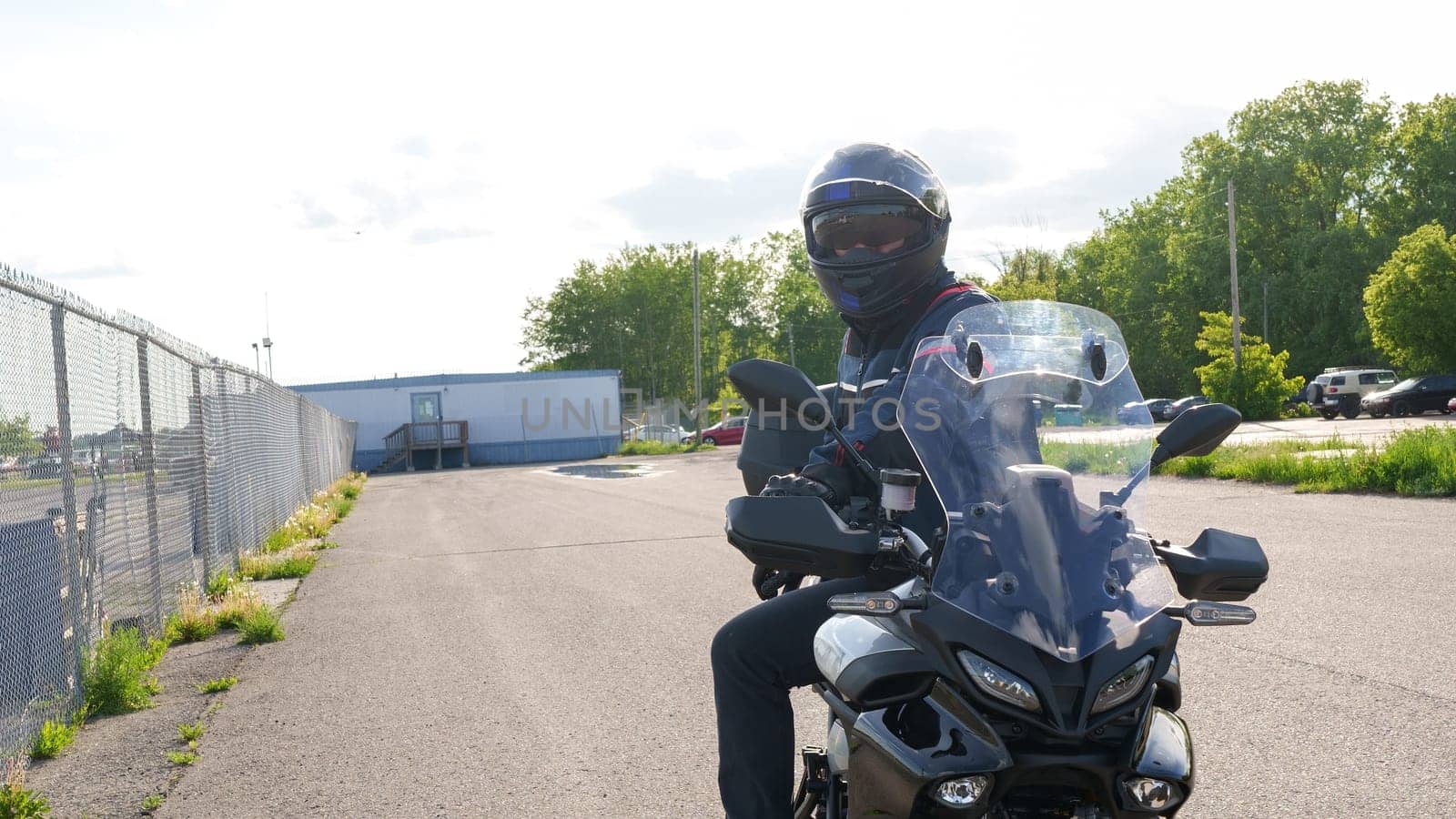 A person confidently sits on a sleek motorcycle in a spacious parking lot. They wear a stylish black jacket and a helmet with a clear visor, while a chainlink fence and lush trees form the backdrop.