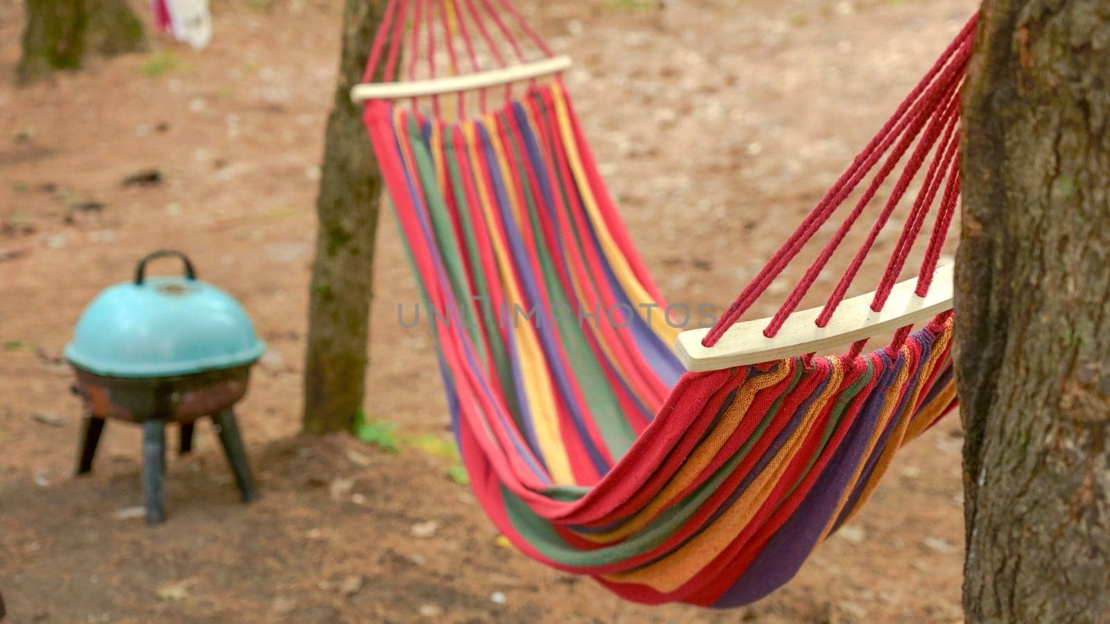 A vibrant, colorful striped hammock is gracefully hanging between two tall trees in a peaceful forest. The hammock remains empty, gently swaying back and forth in the soft breeze, inviting relaxation.