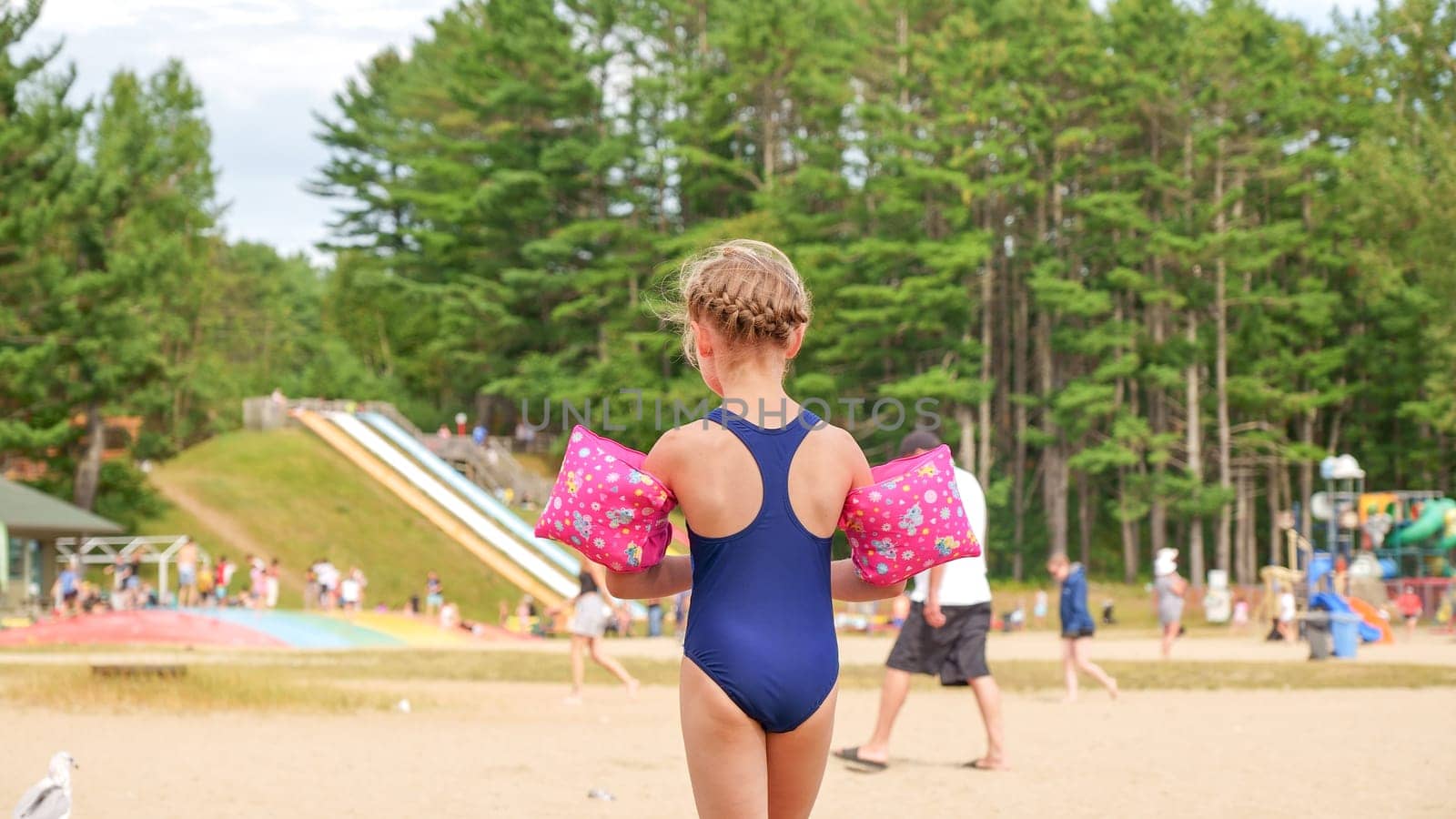 Little girl with pink water wings standing on the sandy beach near the playground on a sunny summer day. The background is blurred, and the girl is wearing a blue one-piece swimsuit.