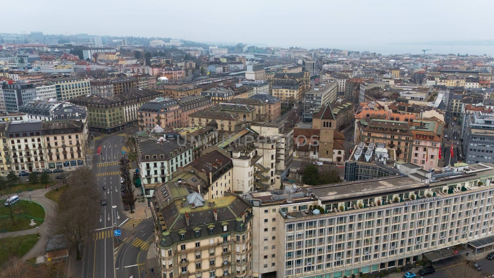 Geneve, Switzerland 04.11.2025 Drone photo of Geneva city center showing historic buildings, a clock tower, hotel, streets, and distant lake under cloudy skies.