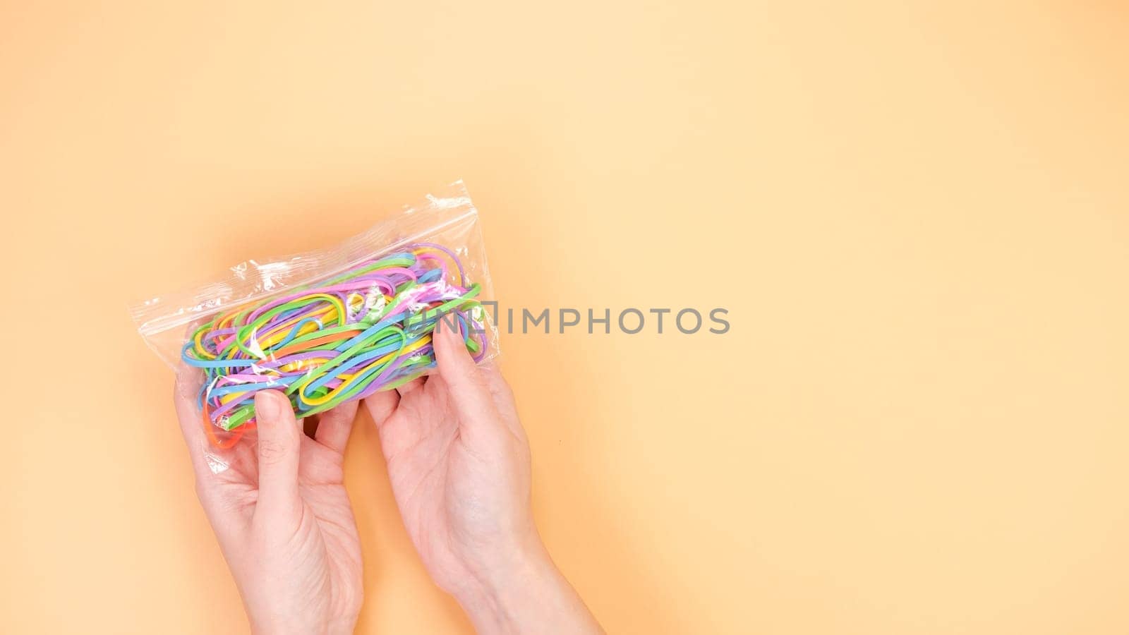 A hand holding a transparent plastic bag filled with colorful rubber bands against a peach background. The rubber bands are in various colors, including pink, blue, green, and yellow.