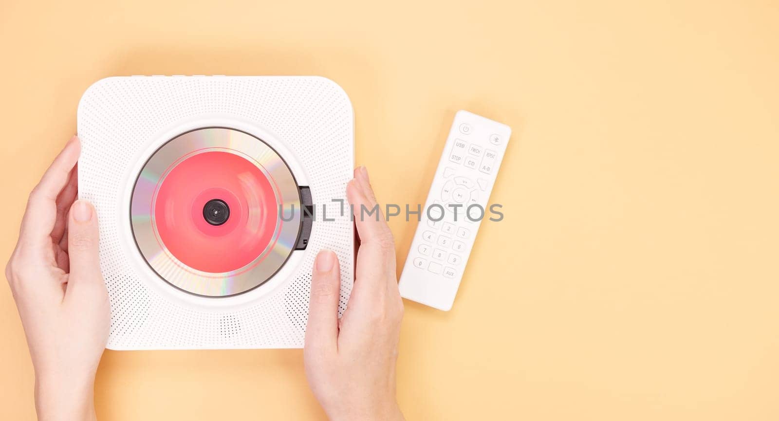 The image shows a white portable CD player featuring a remote control resting on a peach background. Inside the CD player is a vibrant red CD, showcasing its modern design and functionality.