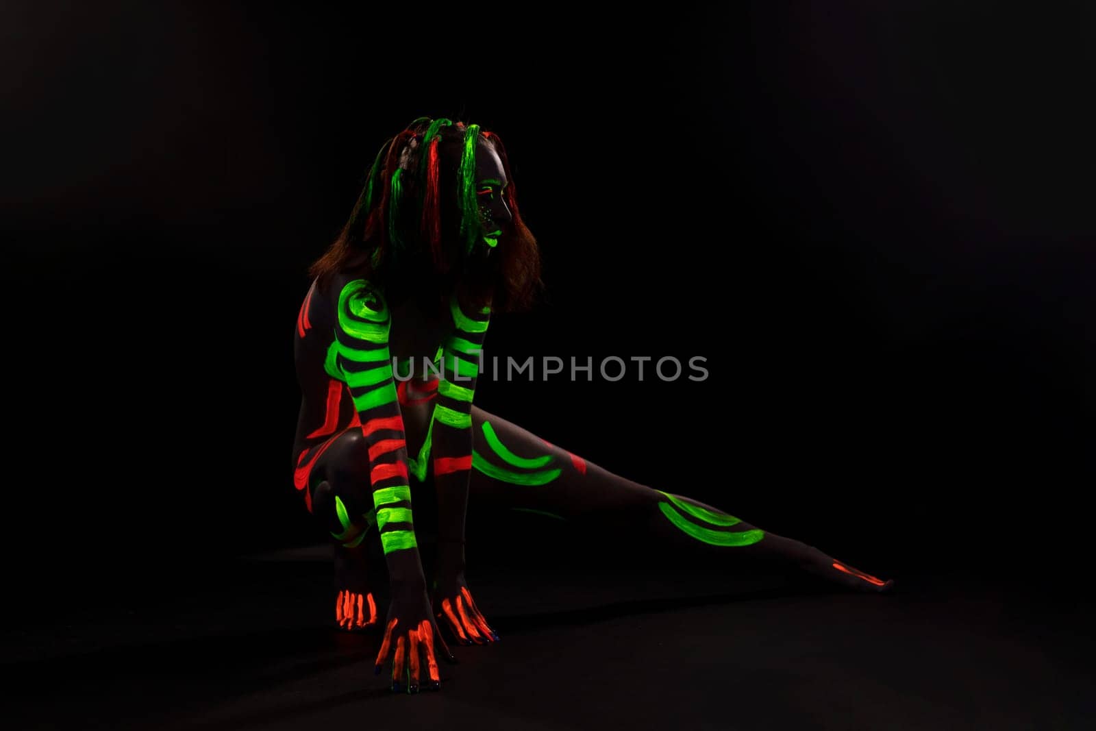 Full body of anonymous female with bright neon red and green fluorescent paints on naked body sitting on haunches in dark studio