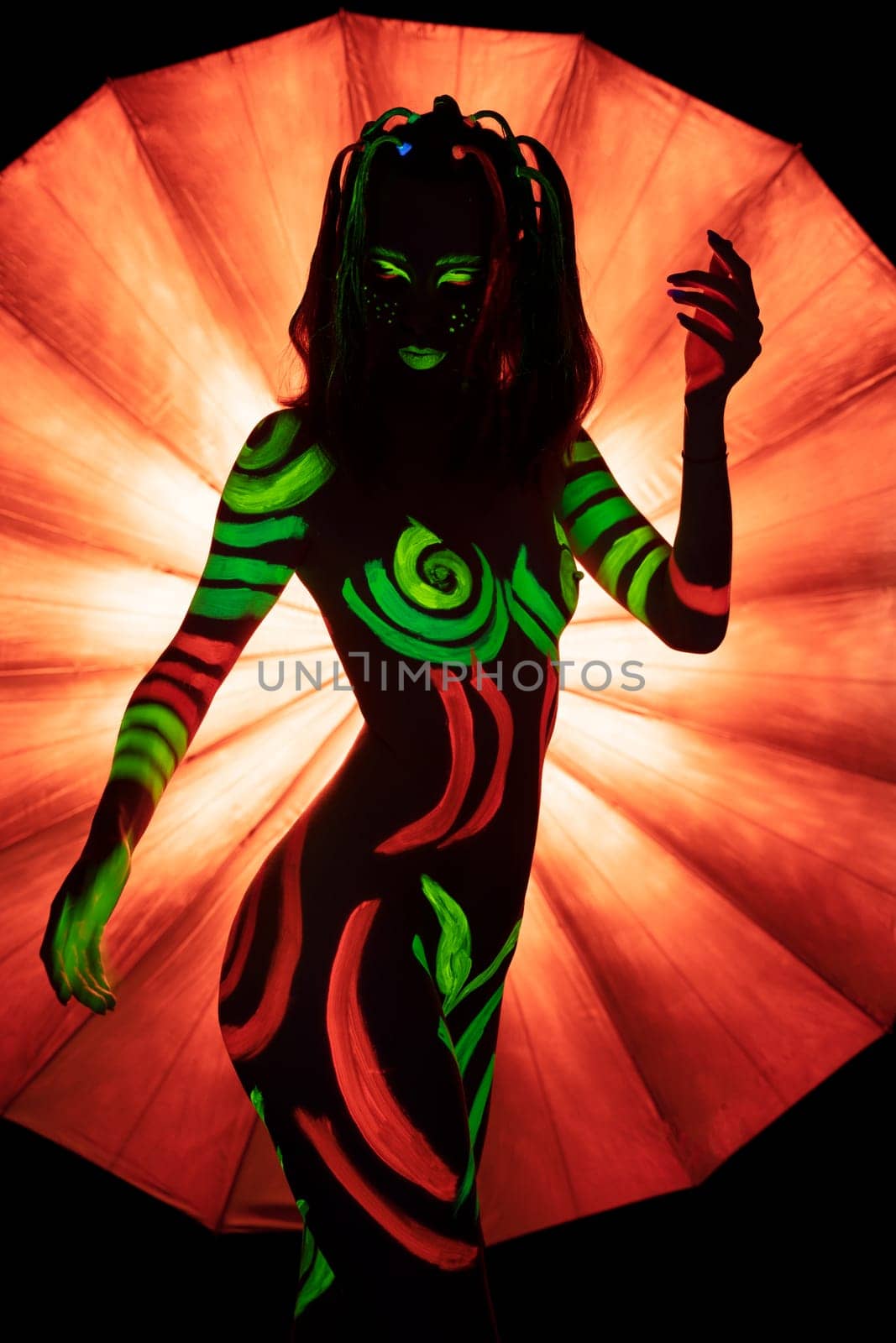 Unrecognizable naked female with bright green and red fluorescent paints on body standing against bright orange umbrella