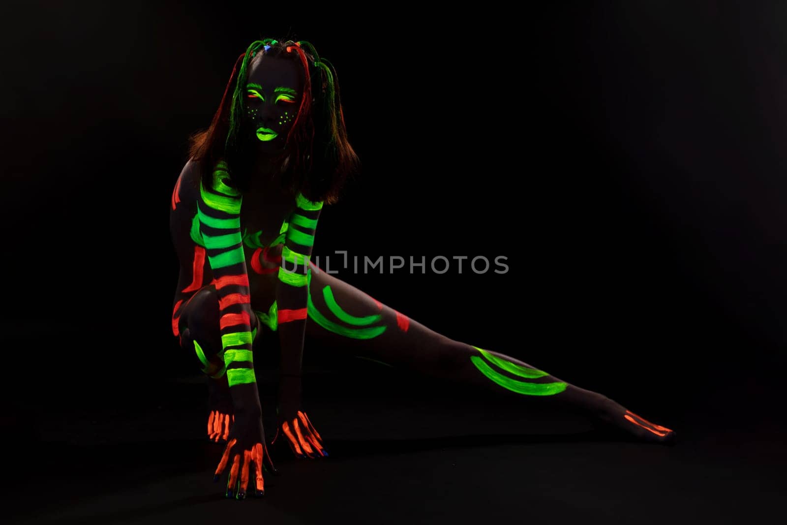 Full body of anonymous female with bright neon red and green fluorescent paints on naked body sitting on haunches in dark studio