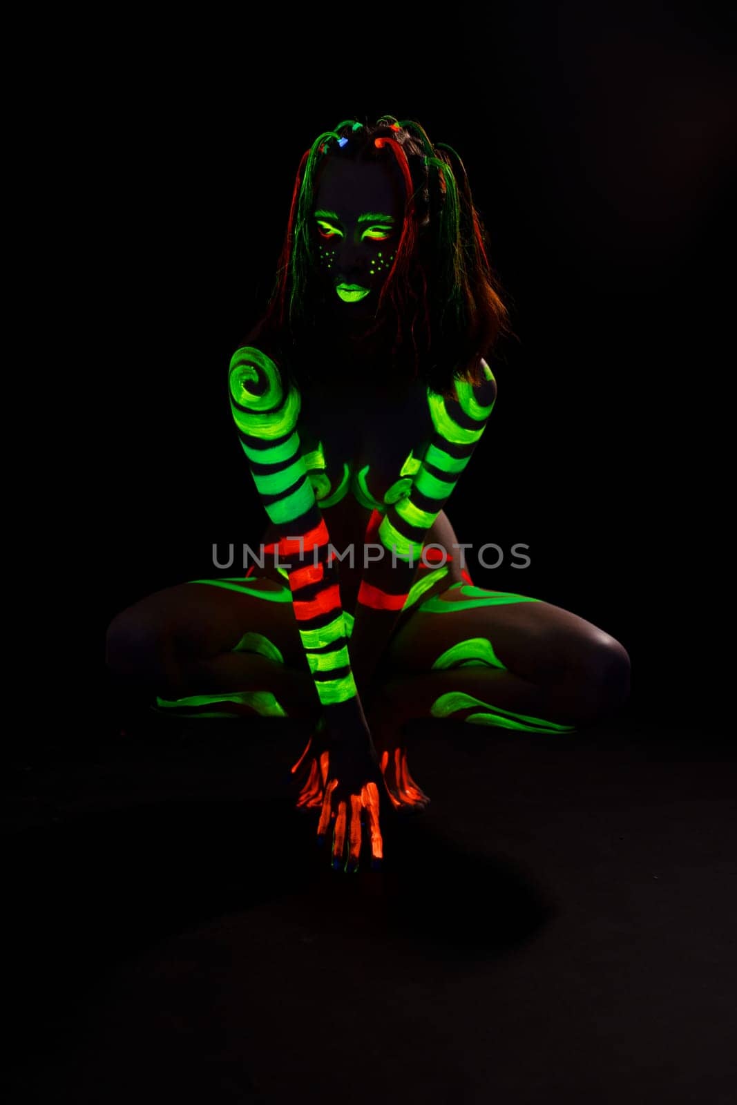 Full body of anonymous female with bright neon red and green fluorescent paints on naked body sitting on haunches in dark studio