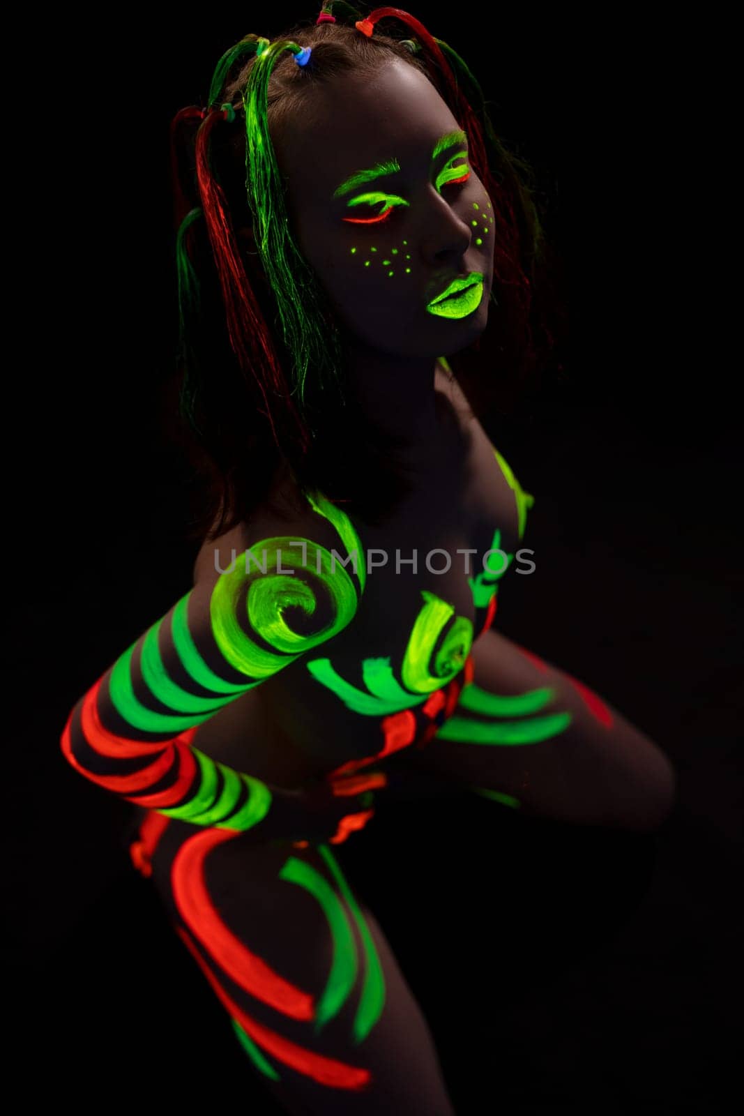 anonymous topless woman with bright neon red and green fluorescent bodyart on body and face stand on knees in dark