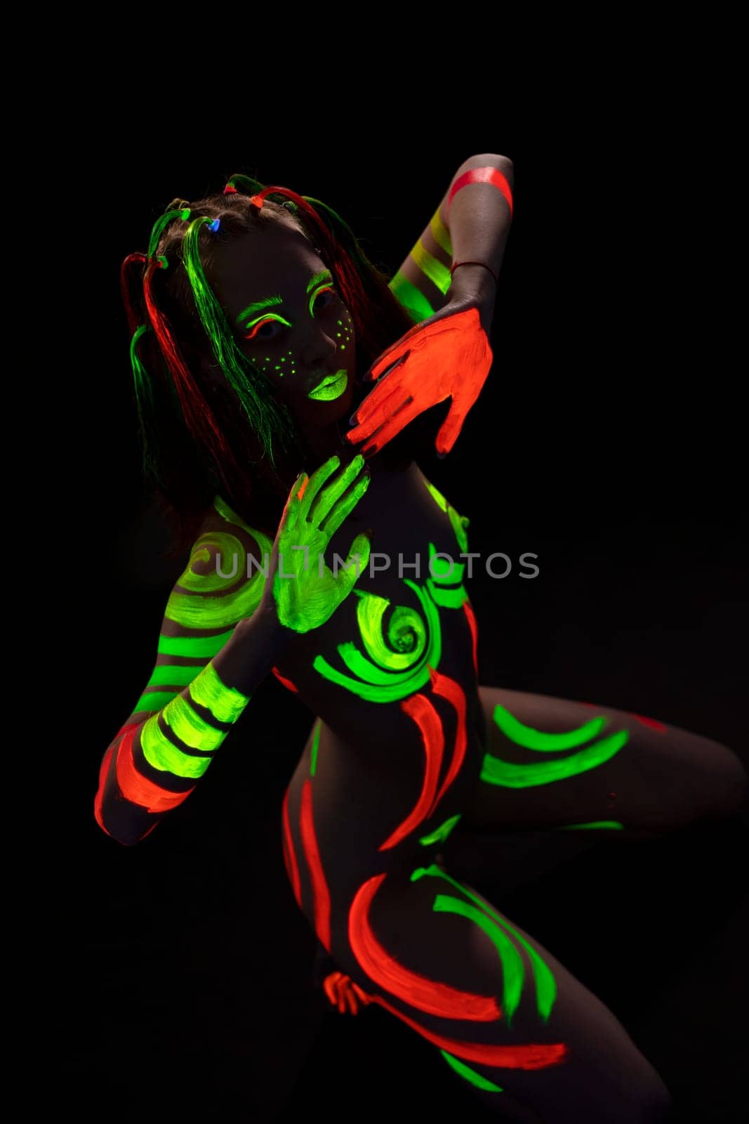 From above of unrecognizable nude lady with creative face and body art made with bright fluorescent paints sitting on haunches in darkness