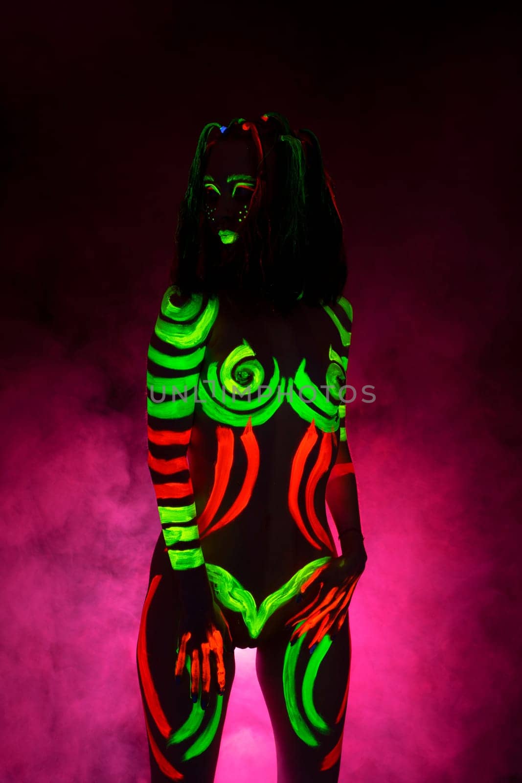 Portrait of unrecognizable female model with bright UV paints on body and face stand on knees in pink smoke