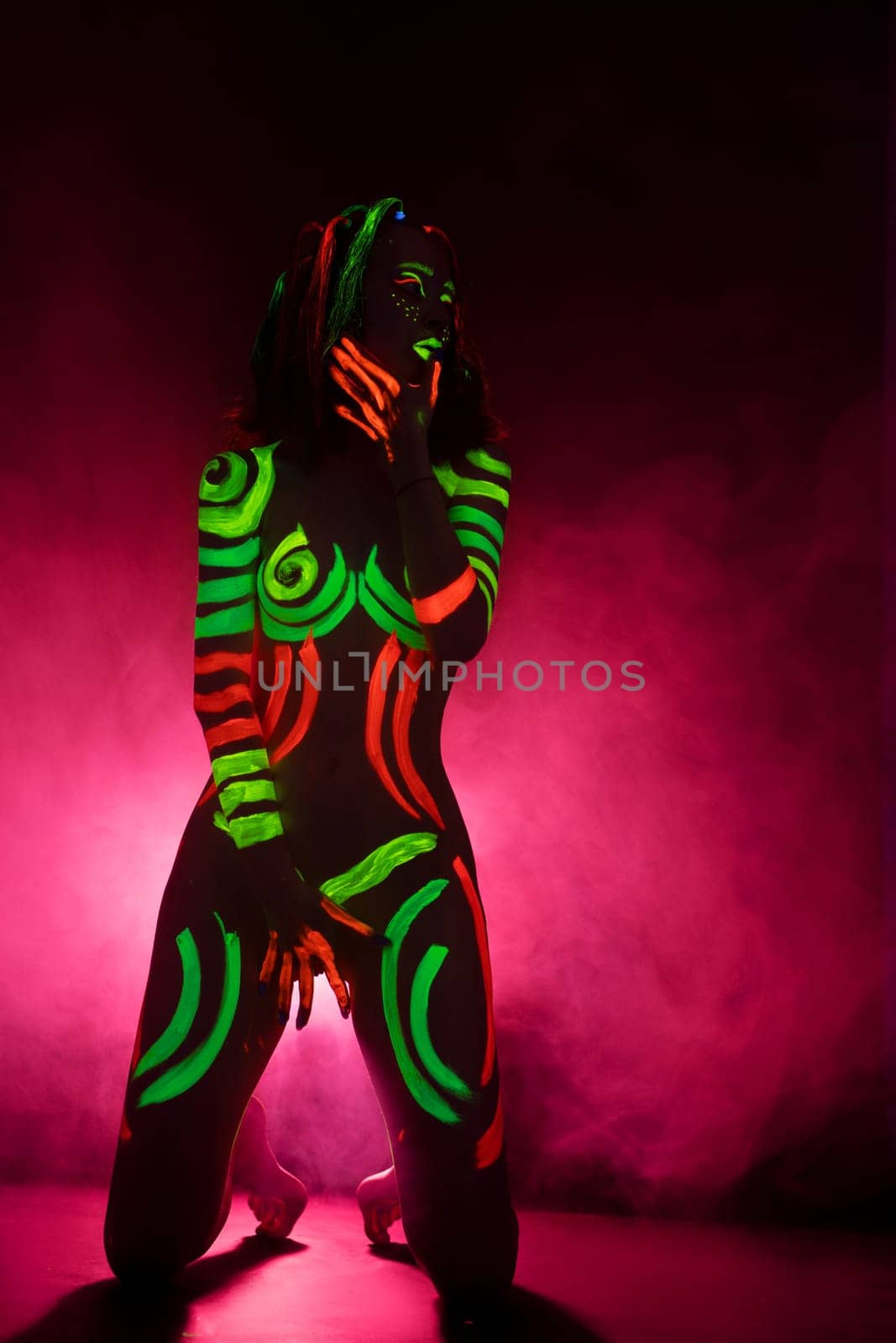 Portrait of unrecognizable female model with bright UV paints on body and face stand on knees in pink smoke