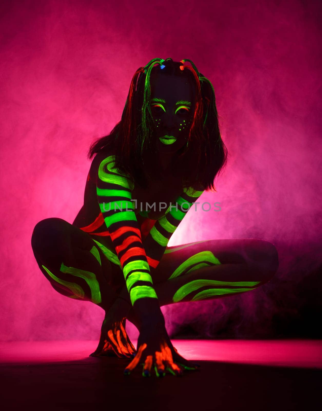 Full length of unrecognizable female model with long hair and bright UV paints on body and face sitting on haunches against pink background