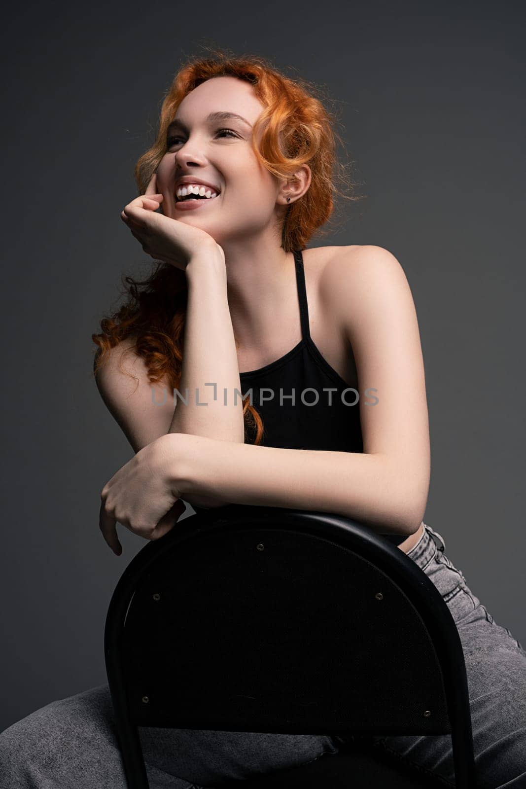 Cheerful young female model with long ginger in casual outfits smiling brightly and looking away while sitting on chair - hand at chin against gray background
