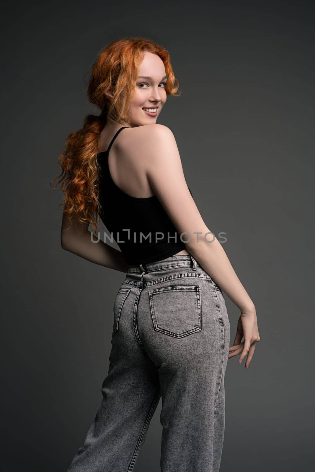 Portrait of young female model in trendy jeans and crop top with red hair stand back in studio with happy emotion - model test