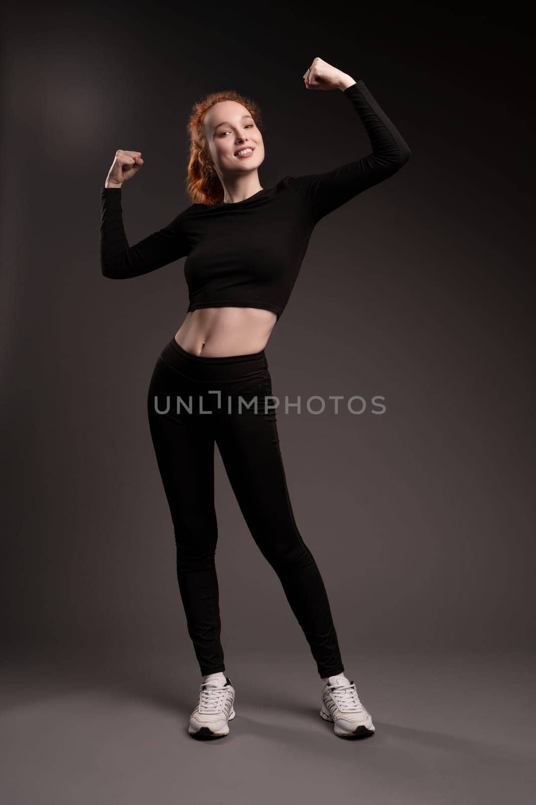 Full body of confident young fit female athlete with long curly red hair in black sportswear show arms and smile on camera