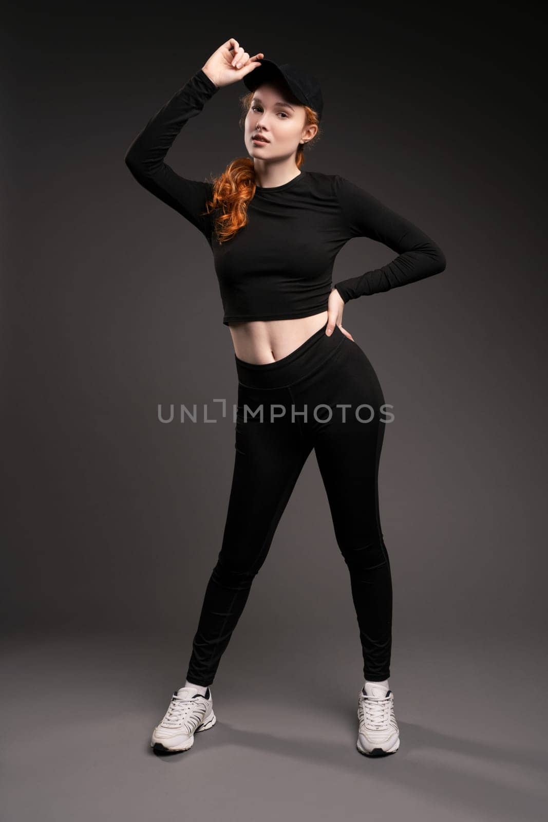 Self assured young female model with curly long red hair in trendy activewear adjusting cap and looking at camera while leaning forward against gray background