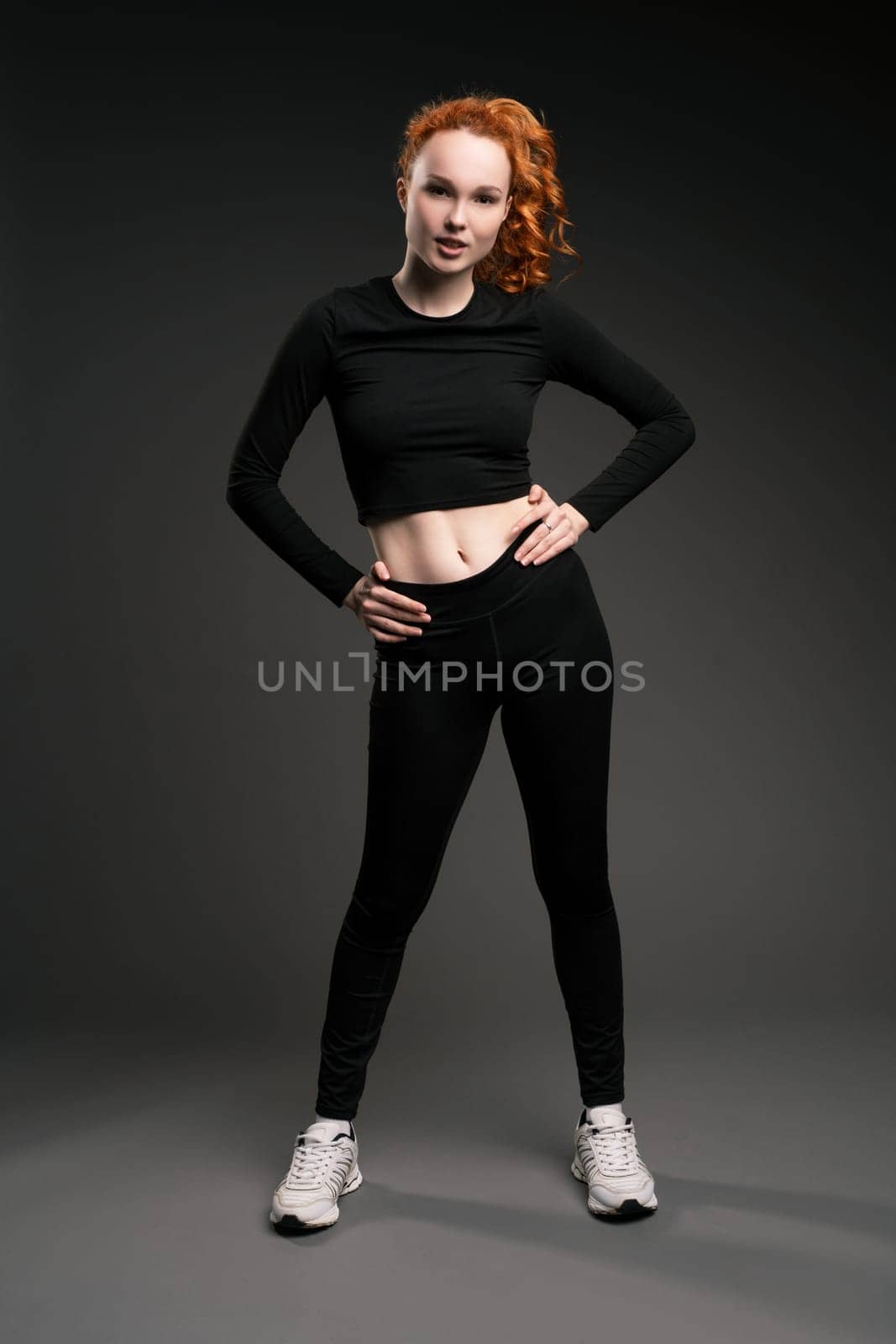 Full body of confident young fit female athlete with long curly red hair in black sportswear and sneakers standing with hands on waist