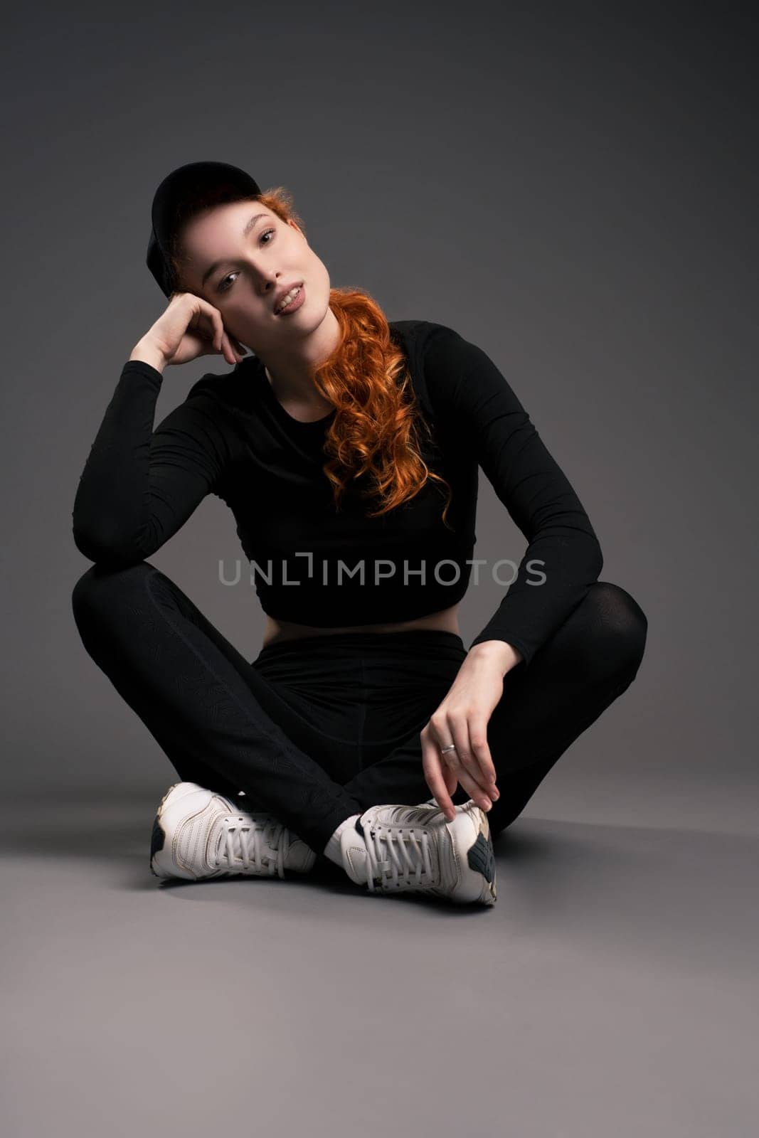 Full body of relaxed young athletic woman with long ginger hair in stylish sportswear and sneakers sitting on floor and looking at camera